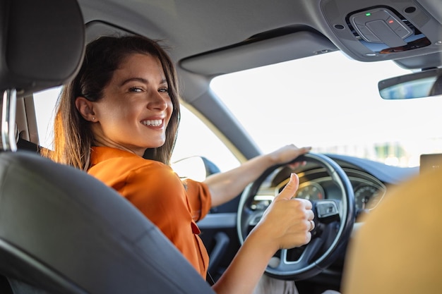 Junge Frau blickt zurück, während sie Auto fährt, und zeigt den Daumen nach oben. Rückansicht einer attraktiven jungen Geschäftsfrau, die ihr beim Autofahren über die Schulter schaut