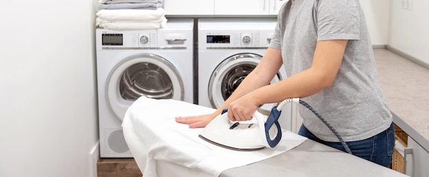 Junge Frau bügelt weißes Hemd an Bord in Waschküche mit Waschmaschine im Hintergrund