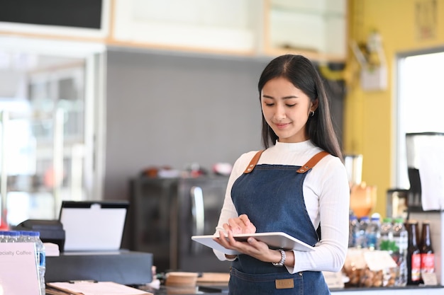 Junge Frau Cafébesitzerin mit Schürze, die digitale Tablette hält, die bereit ist, Bestellungen zu erhalten.