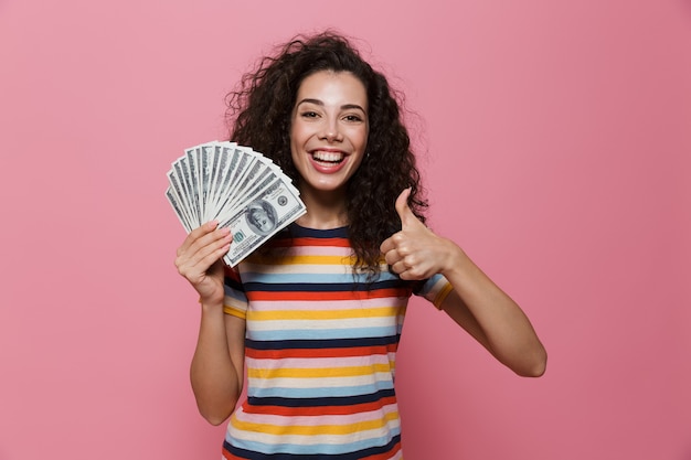 junge Frau der 20er Jahre mit lockigem Haar, die einen Fan von Dollargeld isoliert auf Rosa hält