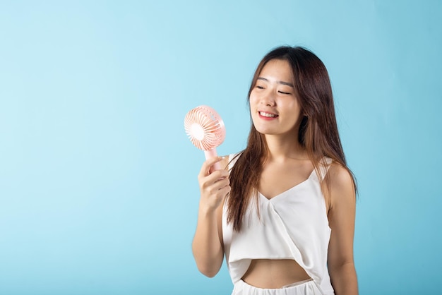 Junge Frau der asiatischen Schönheit, die einen rosafarbenen tragbaren elektrischen Mini-Ventilator in der Nähe ihres Gesichts hält Studioaufnahme isoliert auf blauem Hintergrund, Weibliche Hand hält einen kleinen Plastikventilator, der sie genießt, wenn sie kühlen Wind weht