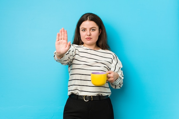 Junge Frau der Übergröße, die eine Teetasse hält, die mit ausgestreckter Hand steht, die Stoppschild zeigt, das Sie verhindert