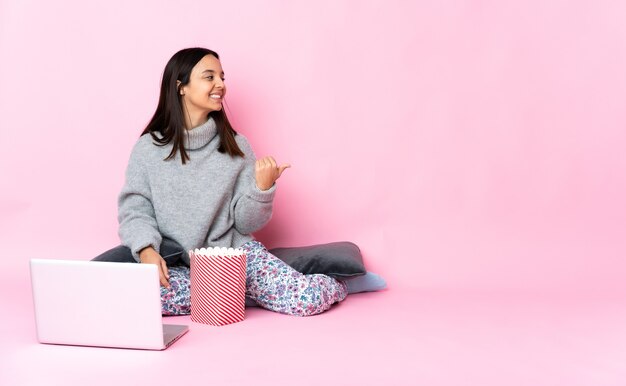 Junge Frau der gemischten Rasse, die Popcorn isst, während sie einen Film auf dem Laptop betrachtet, der zur Seite zeigt, um ein Produkt zu präsentieren