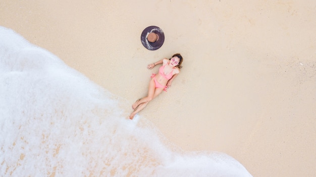 Junge Frau der Vogelperspektivenansicht in einem Bikini, der auf dem Sandstrand und den Wellen liegt