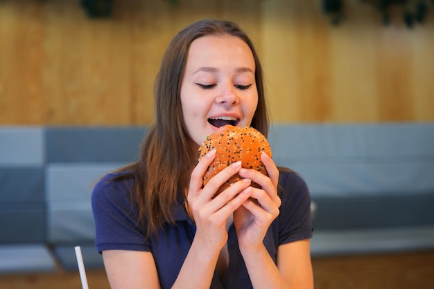 Junge Frau des glücklichen jugendlich Teenagermädchens beißt großen saftigen fetten Burgerhamburger, schnelles ungesundes Essen