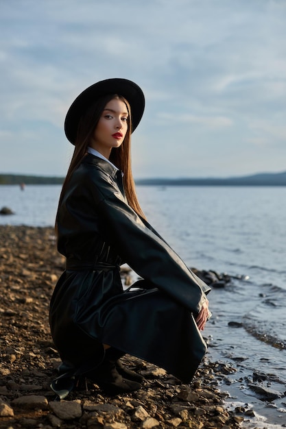 Junge Frau des Porträts bei Sonnenuntergang. Modefrau in schwarzem rundem Hut und ledernem Regenmantel in der Nähe von Seeteich. Schönes Make-up und leuchtend roter Lippenstift auf den Lippen