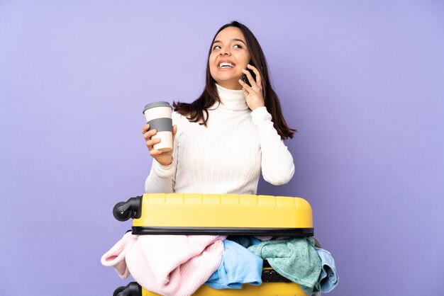 Junge Frau des Reisenden mit einem Koffer voll von Kleidung über der purpurroten Wand, die Kaffee hält, um und ein Mobile wegzunehmen