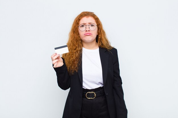 Junge Frau des roten Kopfes, die traurig und weinerlich mit einem unglücklichen Blick fühlt und mit einer negativen und frustrierten Haltung über weißer Wand weint