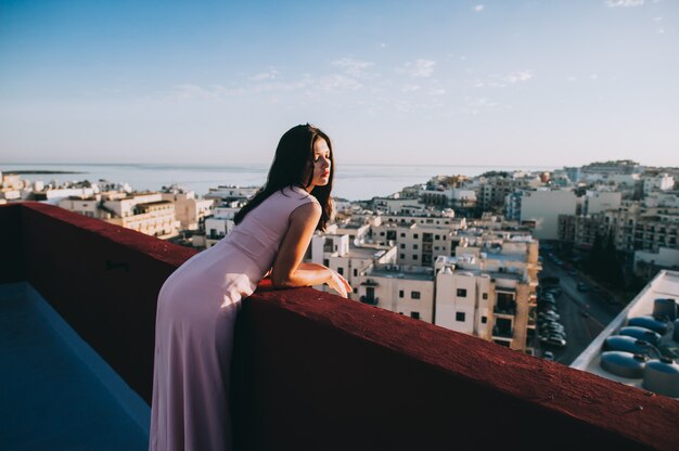 Junge Frau des schönen Brunette in einem weißen Abendkleid. Sonnenuntergang Hintergrund der Stadt einstellen.