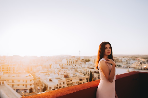 Junge Frau des schönen Brunette in einem weißen Abendkleid. Sonnenuntergang Hintergrund der Stadt einstellen.