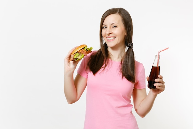 Junge Frau des schönen Spaßes mit den Schwänzen, die Burger, Cola in der Glasflasche lokalisiert auf weißem Hintergrund stehen und halten. Richtige Ernährung oder amerikanisches klassisches Fast Food. Werbefläche mit Kopienraum.