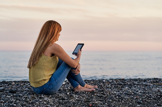 Junge Frau, die allein auf dem Sand am Strand sitzt, während sie ein E-Buch liest. Konzept der Entspannung und des Lesens