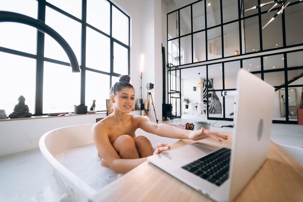 Junge Frau, die am Laptop arbeitet, während sie eine Badewanne nimmt
