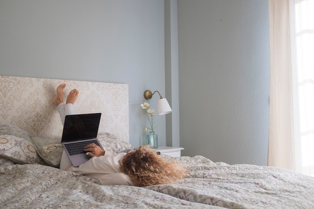 Junge Frau, die am Laptop arbeitet, während sie sich auf dem Bett im Schlafzimmer hinlegt. Geschäftsfrau, die von zu Hause aus arbeitet, während sie sich auf dem Bett entspannt. Faule junge Frau, die auf Laptop schreibt und im Schlafzimmer arbeitet.