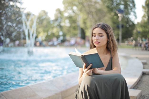junge Frau, die am Rand eines Brunnens in einem Park sitzt, liest ein Buch