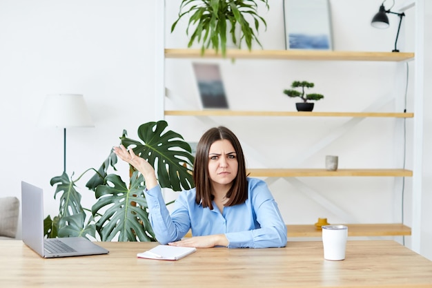 Junge Frau, die am Tisch sitzt und am Laptop arbeitet, verärgerte Frau Das Mädchen breitet ihre Arme aus und ist müde von der Arbeit, sie hat schlechte Laune