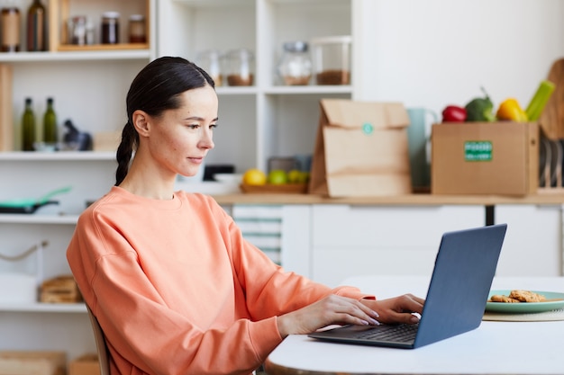 Junge Frau, die am Tisch sitzt und Laptop benutzt, arbeitet er online in der Küche zu Hause