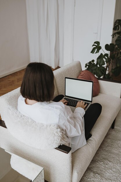 Junge Frau, die an einem Laptop arbeitet, der auf der Couch liegt. Arbeiten Sie zu Hause im Quarantäne-Freiberufler-Konzept