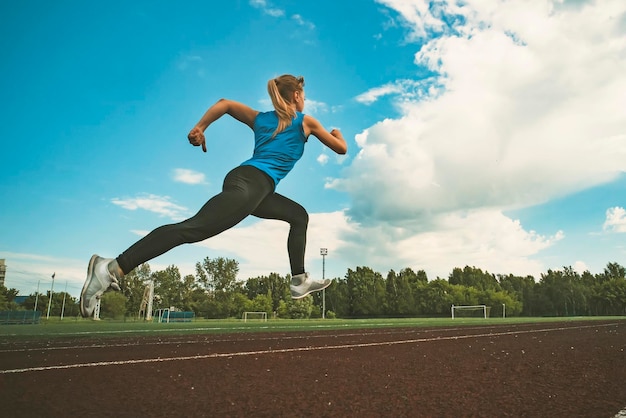 Junge Frau, die an einem sonnigen Abend auf der Stadionstrecke läuft Eine Blondine in einem blauen T-Shirt und schwarzen Leggings läuft durch das Stadion