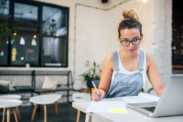 Junge Frau, die Anmerkungen macht und öffentlich mit Laptop studiert.