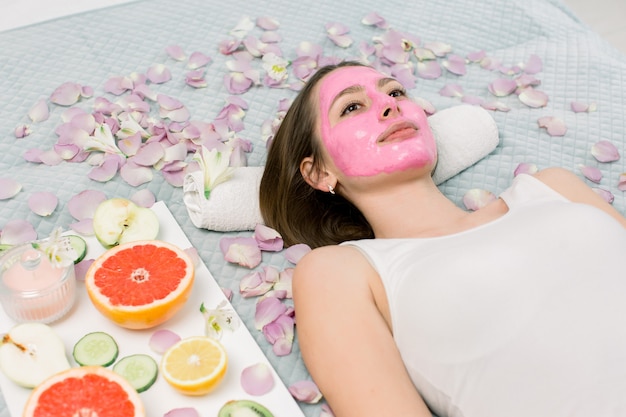 Junge Frau, die auf Bett mit rosa Schlammkosmetikmaske auf Gesicht am Schönheitssalon-Innen entspannt. Blütenblätter, Zitronenscheiben, Grapefruit, Kiwi und Apfel um das Mädchen