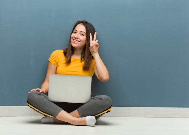 Junge Frau, die auf dem Boden mit einem Laptopspaß sitzt und glücklich ist, eine Geste des Sieges tuend