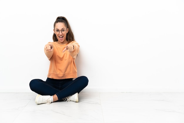 Junge Frau, die auf dem Boden sitzt, überrascht und nach vorne zeigend