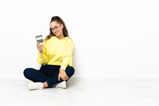 Junge Frau, die auf dem Boden sitzt und einen Kaffee zum Mitnehmen hält, der viel lächelt