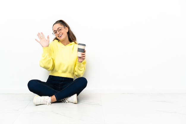 Junge Frau, die auf dem Boden sitzt und einen Kaffee zum Mitnehmen mit der Hand mit glücklichem Ausdruck salutiert