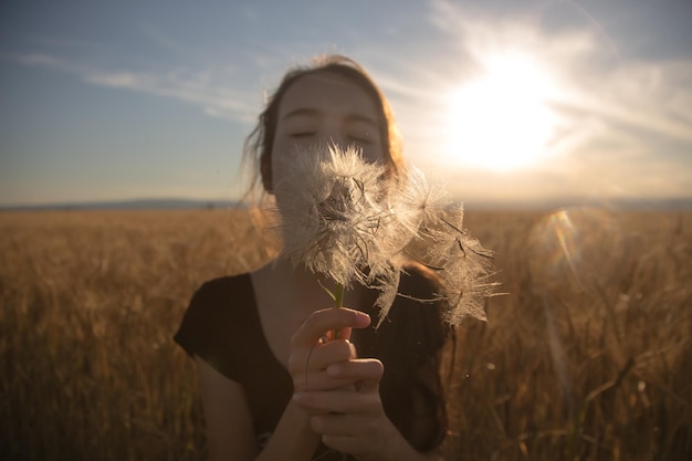 Junge Frau, die auf dem Feld Dandelion gegen den Himmel bläst