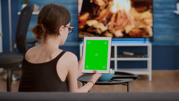 Junge Frau, die auf dem Sofa sitzt und ein vertikales digitales Tablet mit grünem Bildschirm hält, das Online-Social-Media-Inhalte im Wohnzimmer anschaut. Mädchen mit Touchscreen-Gerät mit Chroma-Key im Online-Videoanruf.