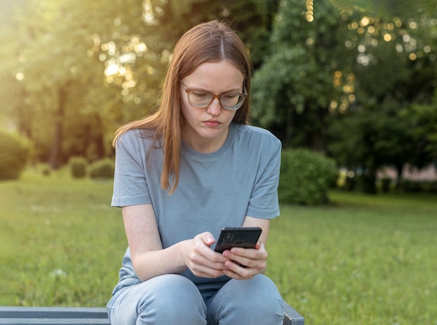 Junge Frau, die auf der Bank sitzt und das Telefon ernste ängstliche Person benutzt, die auf dem Smartphone in der Natur schaut