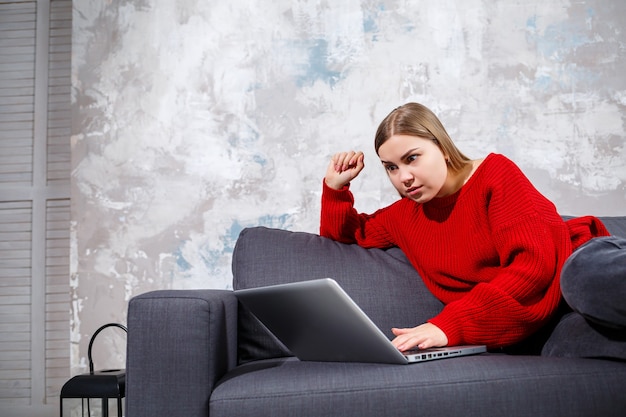 Junge Frau, die auf der Couch sitzt und den Laptopbildschirm betrachtet. Eine motivierte, zu Hause arbeitende Studentin, die online an einem Computer arbeitet.