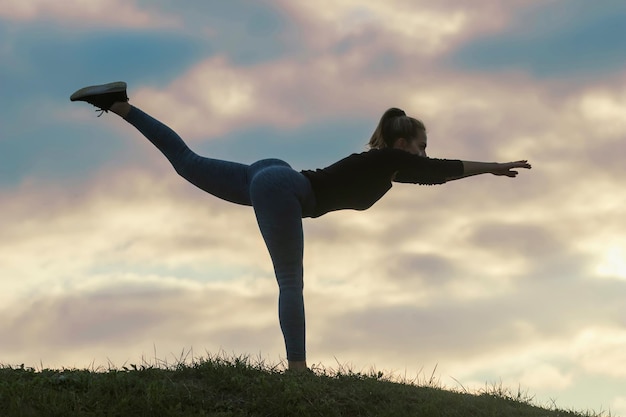 Junge Frau, die auf einem Bein steht und auf dem Gras-Morgen-Training trainiert, schöner Sonnenaufgang