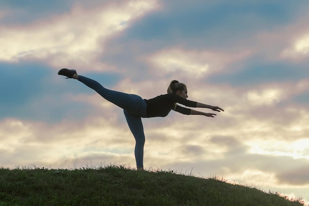 Junge Frau, die auf einem Bein steht und auf dem Gras-Morgen-Training trainiert, schöner Sonnenaufgang