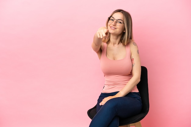 Junge Frau, die auf einem Stuhl über isoliertem rosa Hintergrund sitzt, zeigt mit einem selbstbewussten Ausdruck mit dem Finger auf dich