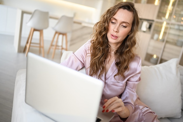 Junge Frau, die auf einem weißen Sofa im Wohnzimmer der skandinavischen Artwohnzimmerküche sitzt und an einem Laptop arbeitet.