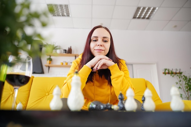 Junge Frau, die auf gelbem Sofa sitzt und im Zimmer Schach spielt Weibliches Spielen im logischen Brettspiel mit sich selbst