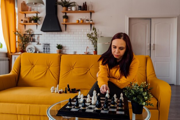 Junge Frau, die auf gelbem Sofa sitzt und im Zimmer Schach spielt Weibliches Spielen im logischen Brettspiel mit sich selbst