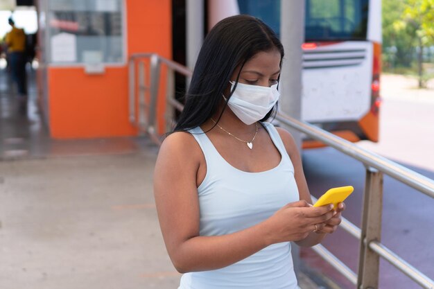 Junge Frau, die auf ihr Telefon schaut, während sie auf ihren Bus am Bahnhof wartet. Sie trägt aufgrund der COVID-19-Pandemie eine Maske.