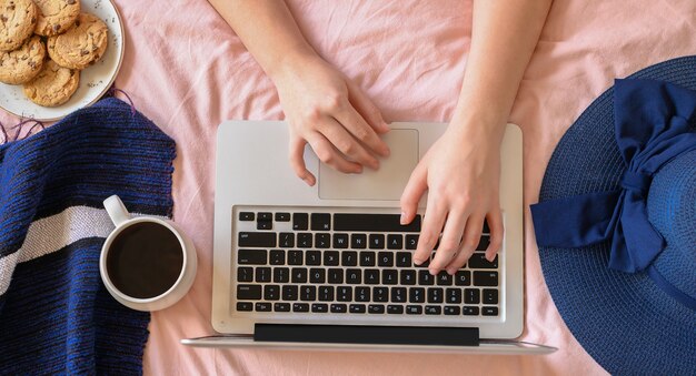 Junge Frau, die auf Laptop im Bett tippt. Lifestyle-Heldenkopf mit Blick von oben mit Tasse Kaffeehut. Freiberufler oder Modeblogger zu Hause Arbeitsbereich.