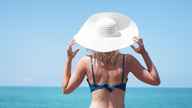 Junge Frau, die auf Sand nahe Meer steht und einen weißen Hut hält