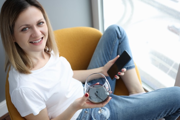 Junge Frau, die auf Sessel mit Glas Rotwein und Telefon sitzt