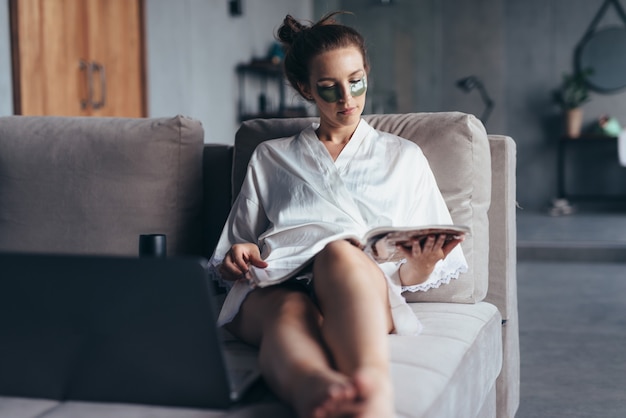 Junge Frau, die auf Sofa mit Flecken unter Augen und Lesemagazin sitzt.