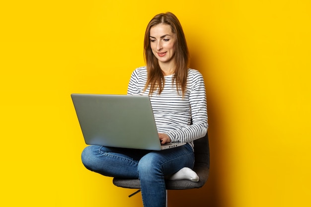 Junge Frau, die auf Stuhl sitzt und Laptop auf lokalisiertem Gelb hält.