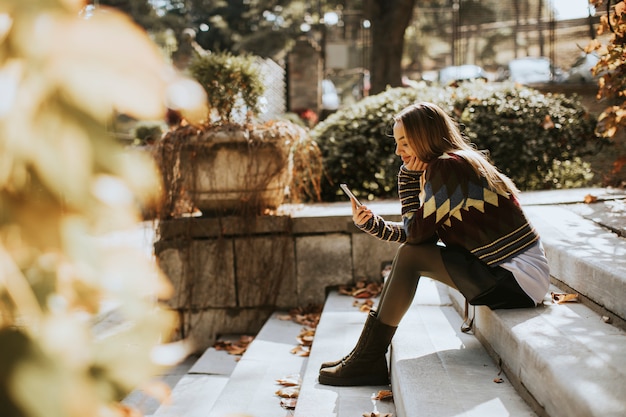 Junge Frau, die auf Treppen mit Handy im Herbstpark sitzt