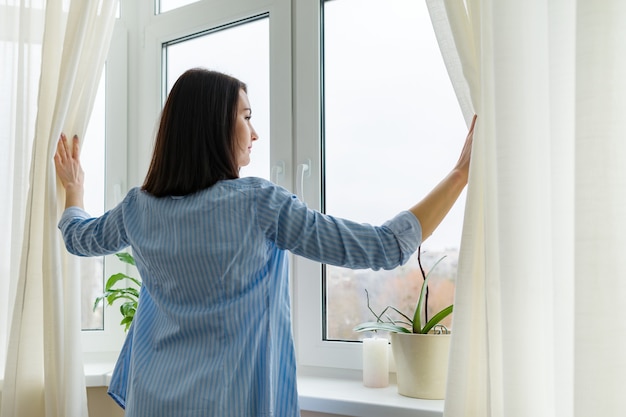 Junge Frau, die aus dem Fenster schaut, bewölkter Wintertag, Frau im blauen Hemd, das Vorhänge öffnet