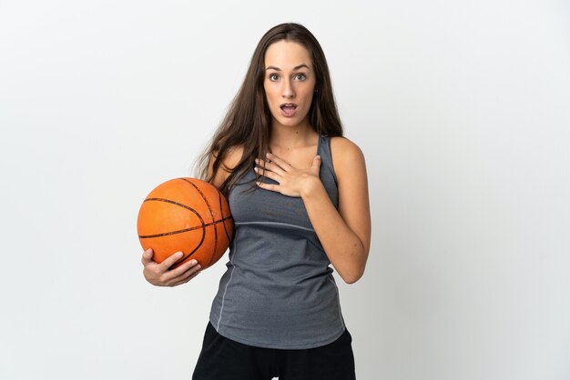 Junge Frau, die Basketball über isoliertem weißem Hintergrund spielt, überrascht und schockiert, während sie nach rechts schaut