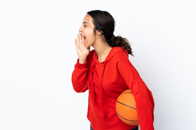 Junge Frau, die Basketball über isoliertem weißem Hintergrund spielt und mit weit geöffnetem Mund zur Seite schreit