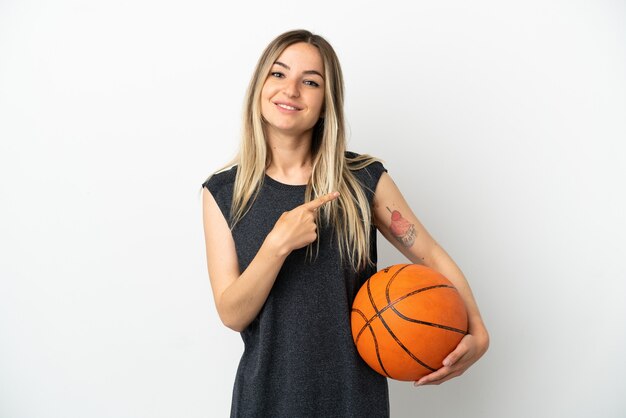 Junge Frau, die Basketball über isolierter weißer Wand spielt, die auf die Seite zeigt, um ein Produkt zu präsentieren?